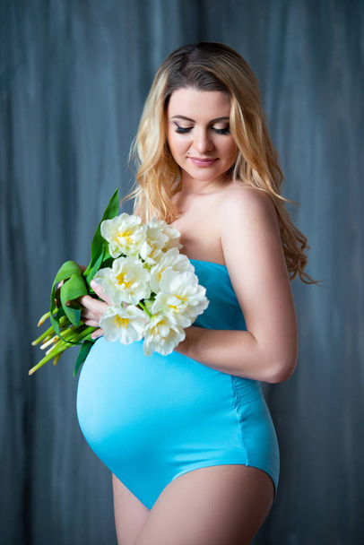 Beautiful young pregnant blonde woman holding flowers white tulips in her hands. Spring mood, freshness, women's Day, health and beautiful pregnancy - Фото, изображение