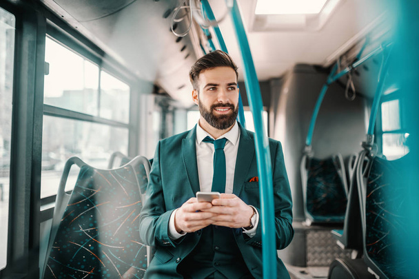 Smiling ambitious bearded businessman in turquoise suit sitting in public bus and using smart phone. If you want to fly, give up everything that weights you down. - Foto, imagen