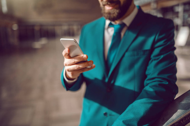 Close up of bearded smiling Caucasian businessman using smart phone while waiting on airport. Business trip concept. Sell the problem you solve, not the product. - Foto, Imagen