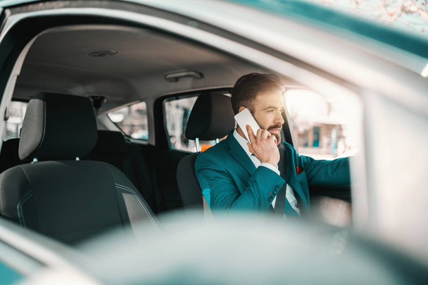 Fiatal fehér sikeres szakállas üzletember türkiz öltönyben ül a kocsiban és okos telefont használ. Próbálj egy kicsit keményebb lenni, hogy egy kicsit jobb legyél.. - Fotó, kép