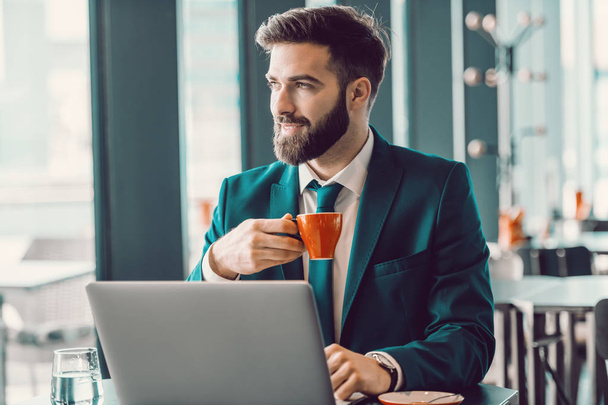 Homem de negócios elegante caucasiano sorrindo bebendo café no café, usando laptop e olhando pela janela. Empurra-te, porque ninguém o vai fazer por ti.
. - Foto, Imagem