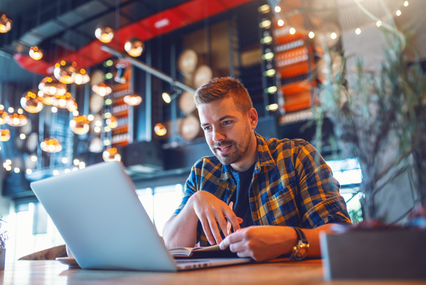 Sonriente blogger masculino caucásico en camisa a cuadros con agenda y mirando a la computadora portátil mientras está sentado en la cafetería
. - Foto, imagen