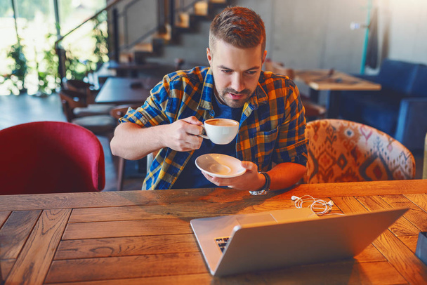 Junge kaukasische Bloggerin im karierten Hemd trinkt Kaffee und schaut auf Laptop, während sie im Café sitzt. - Foto, Bild