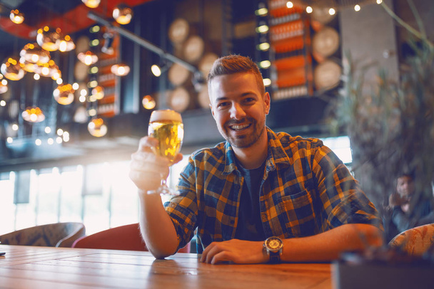 Sonriente caucásico guapo hombre en camisa a cuadros sentado en el bar y sosteniendo el vaso de cerveza mientras mira a la cámara
. - Foto, Imagen