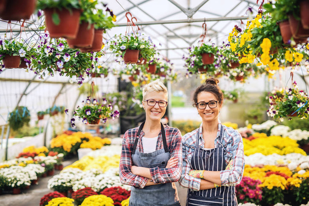 Dos floristas caucásicos en delantales de pie en invernadero con los brazos cruzados rodeados de flores
. - Foto, Imagen