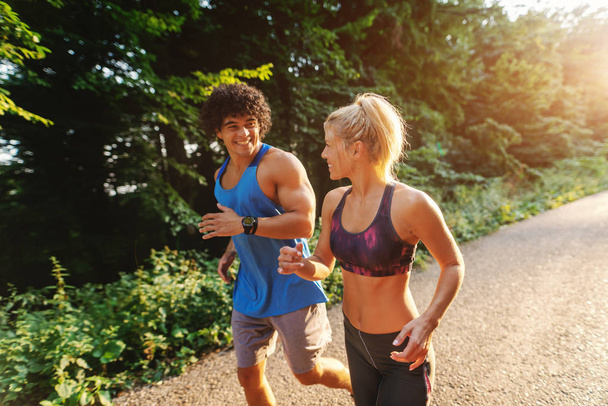 Vista lateral de la linda pareja deportiva corriendo en la naturaleza en el soleado día de verano
. - Foto, Imagen