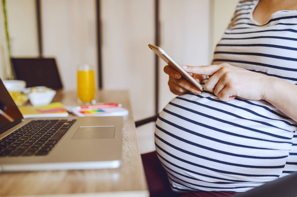 テーブルに座っている間、若い白人妊婦はスマートフォンを使用しています。ジュースとテーブルのラップトップとガラスで。妊娠中の女性の概念. - 写真・画像