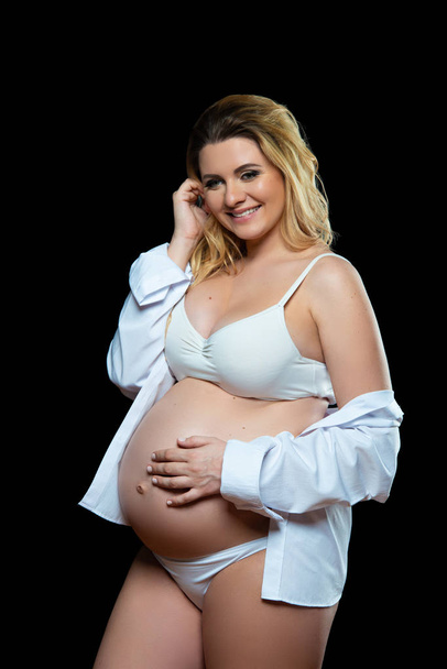 Beautiful smiling blonde pregnant woman in white clothes on a black background. Women's health and happy pregnancy - Foto, Imagen