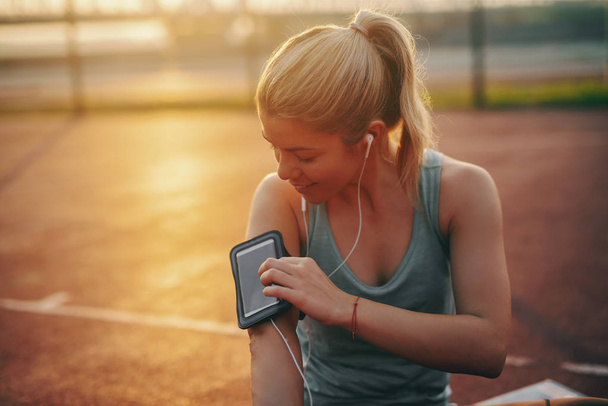 Uśmiechnięty piękny kaukaski sportowy kobieta blondynka z etui na telefon wokół ramienia siedzi na boisku i włączenie muzyki na inteligentny telefon. Inteligentny telefon w etui. Koncepcja porannej fitness. - Zdjęcie, obraz
