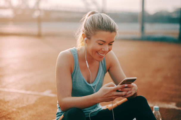 Cute smiling Caucasian blonde sporty woman sitting on the court with earphones in ears and using smart phone. - Photo, Image