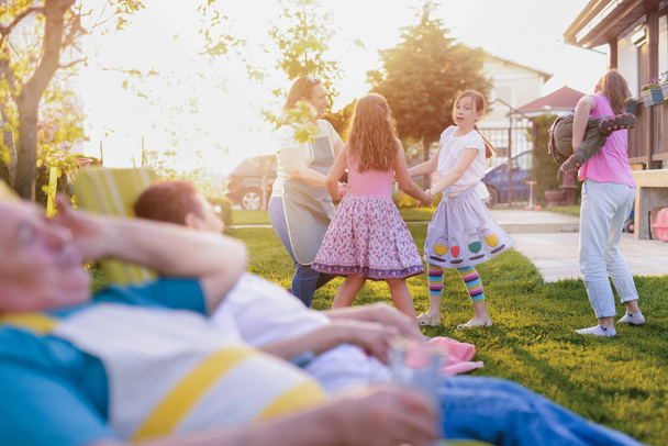 Gran familia feliz en su patio trasero divertirse y pasar tiempo juntos. Abuela y abuelo tumbados en sillas y viendo a sus nietas jugar. - Foto, Imagen
