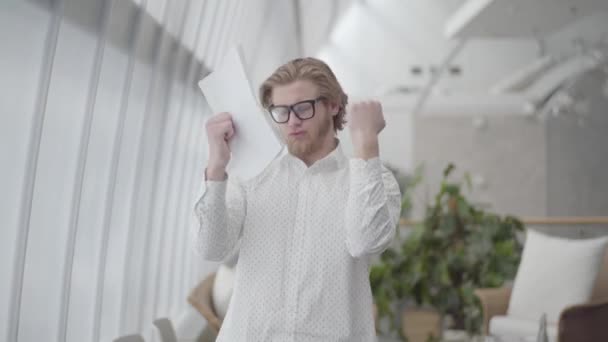 Blond happy man in glasses dancing in a light comfortable office holding papers in hands. The boss throws documents up. Handsome businessman has good news. Workplace. Office space. - Metraje, vídeo