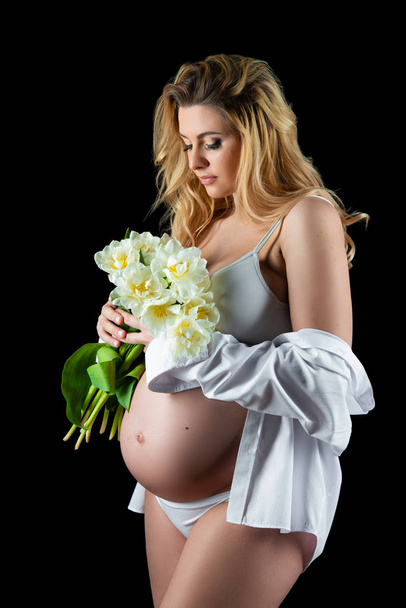 Beautiful blonde woman with a pregnant tummy wearing a white shirt and holding flowers white tulips in her hands. Girl posing on a black background - Photo, image