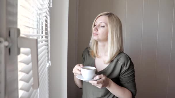 Young sad thinkful beautiful blond woman standing near the window with blinds in the morning and drinking coffee - Imágenes, Vídeo