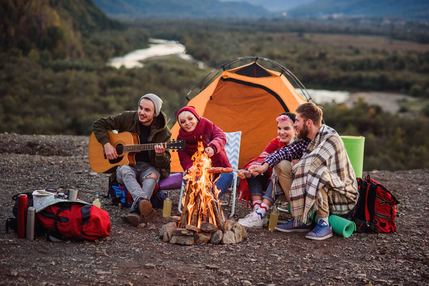 Happy friends having a picnic at the mountains, they are chatting, drinking energy drinks, playing on guitar and bake sausages on campfire. Concept of happiness, youth and enjoyment of life. - Foto, Bild