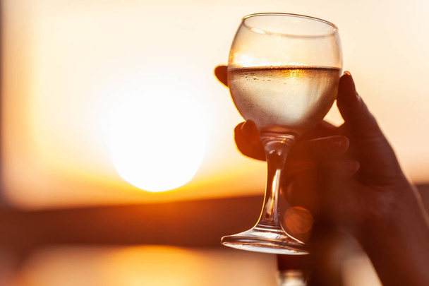 Een veld glas koude witte wijn in een dames hand, als goud als zonsondergang op de achtergrond - Foto, afbeelding