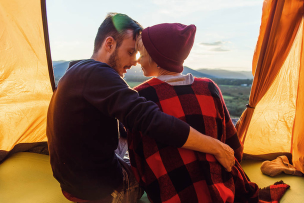 Young man and his girlfriend sitting in the tent, leaning against each other on the background of the mountains at sunset. View from back. - Фото, зображення
