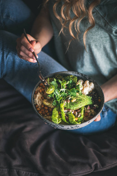 Healthy dinner or lunch. Curly woman in t-shirt and jeans sitting and eating vegan superbowl or Buddha bowl with hummus, vegetable, fresh salad, beans, couscous and avocado. Clean eating food concept - Fotó, kép