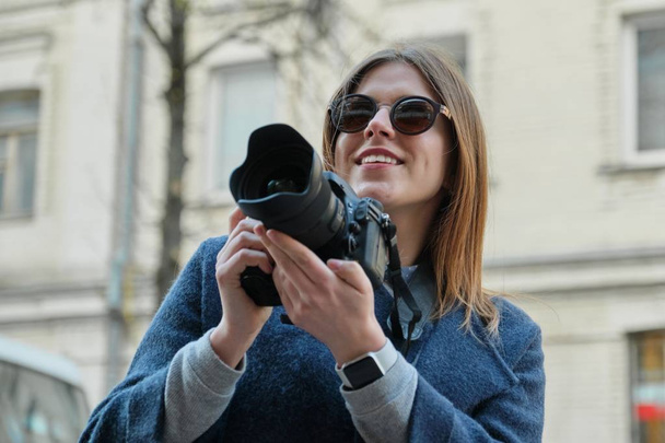Young beautiful girl with camera in the spring city, girl tourist taking photographs on city street - Fotó, kép