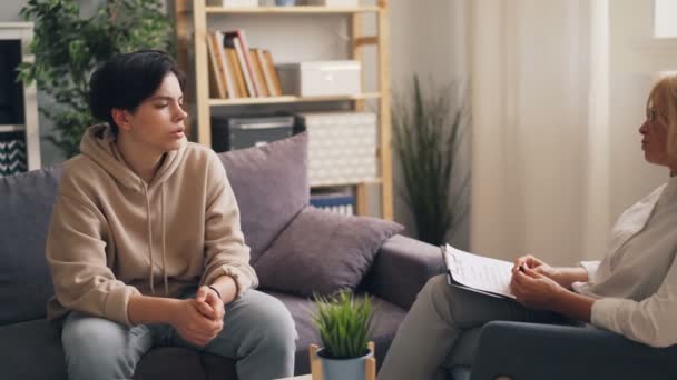 Sad teenage boy talking to female psychotherapist sitting on sofa in office - Filmati, video