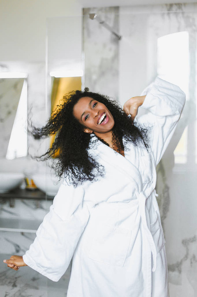 Young afro american cute woman dancing in bathroom and wearing white bathrobe. - Fotoğraf, Görsel
