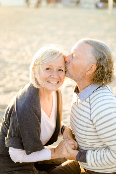 Szczęśliwy starszy mąż całuje starszą żonę i siedzi na piaszczystej plaży. - Zdjęcie, obraz