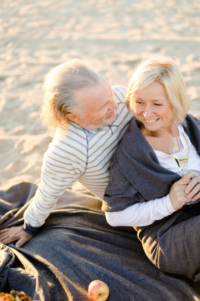 Senior husband hugging smiling wife sitting on plaid with champagne on sand beach. - Photo, Image