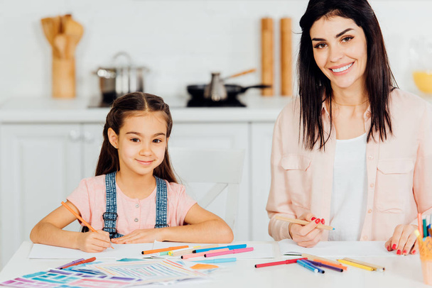 cheerful mother and cute daughter looking at camera and smiling while holding color pencils  - Foto, Bild