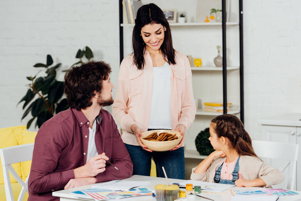 cheerful mother holding bowl with cookies near husband and daughter  - Photo, Image