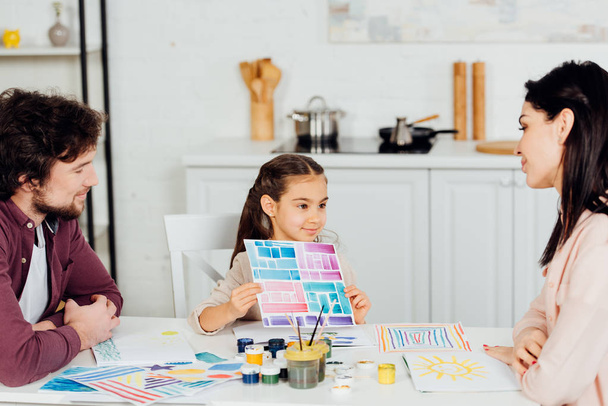 niño alegre sosteniendo papel con rayas de colores cerca de los padres en casa
  - Foto, imagen
