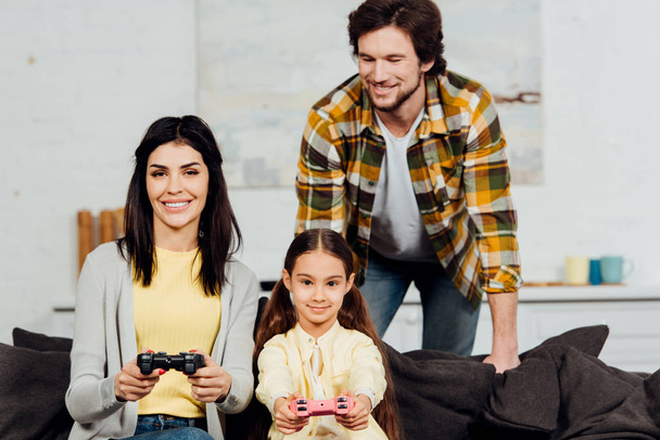 happy man looking at cheerful wife and kid playing video game at home  - Valokuva, kuva