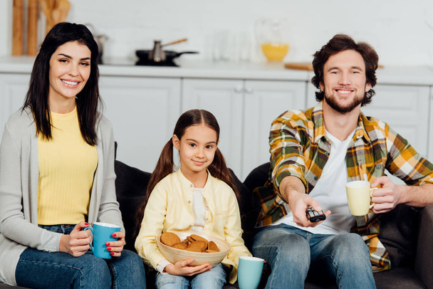 happy man holding remote controller and holding cup near family  - Foto, afbeelding