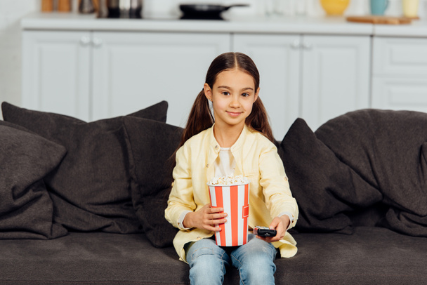 cheerful kid holding remote controller while sitting on sofa with bucket of popcorn  - Foto, afbeelding