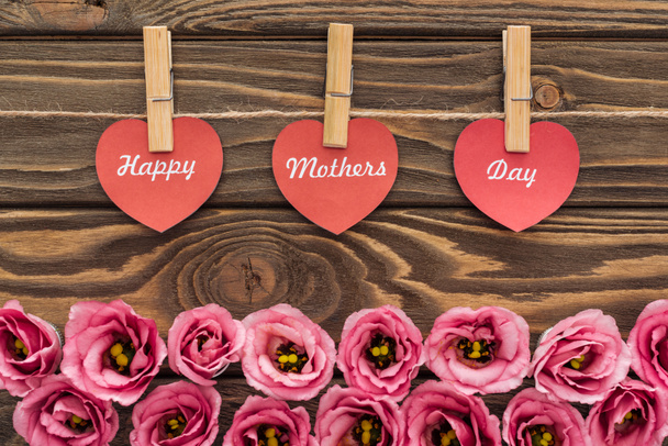 vista dall'alto di fiori di eustoma rosa, mollette di vestiti e cuori di carta rossa con scritte di giorno di madri felici su tavolo di legno
 - Foto, immagini