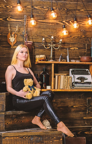 Girl in black clothes holds teddy bear toy in hand, wooden interior on background. Woman on dreamy face relaxing in wooden interior. Lady blonde enjoy leisure with teddy bear. Rest and relax concept - Fotoğraf, Görsel