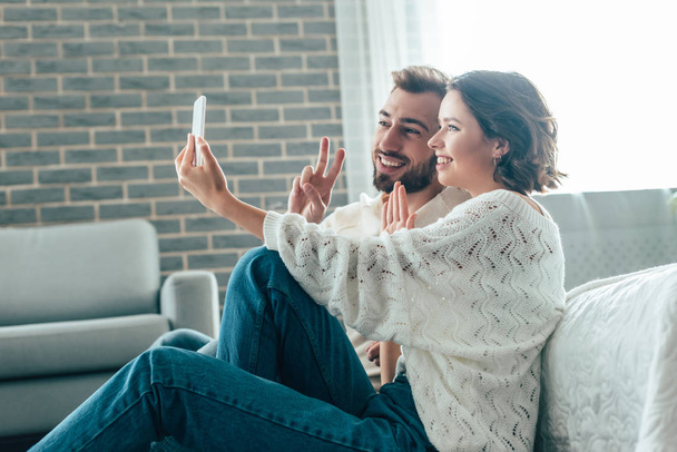 happy woman waving hand while taking selfie with man showing peace sign at home  - Photo, image