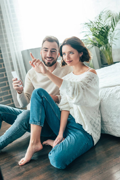 attractive brunette woman pointing with finger while sitting with handsome man holding smartphone at home  - Foto, afbeelding