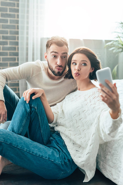 selective focus of attractive woman and handsome man with duck faces taking selfie at home  - Фото, изображение