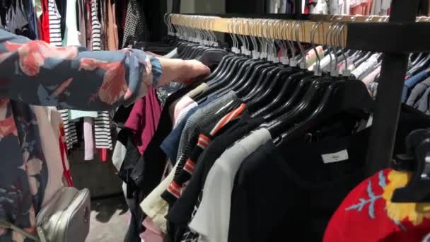 Ψώνια σε ένα κατάστημα ρούχων. Η γυναίκα διαλέγει ρούχα που κρέμονται από κρεμάστρες.. - Πλάνα, βίντεο