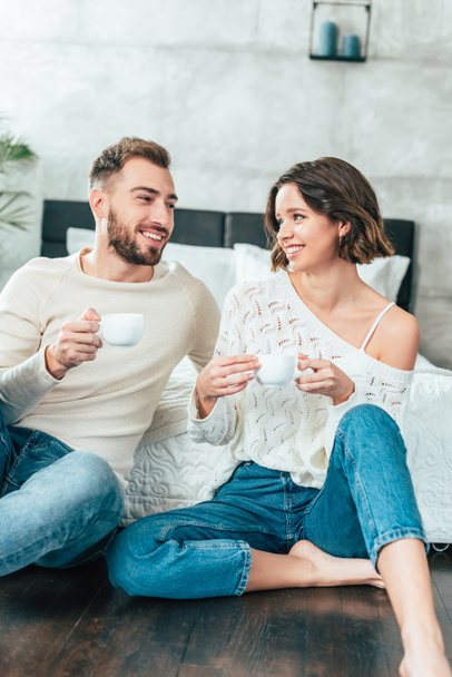 happy man and woman sitting on floor and holding cups with drinks - Foto, Imagem