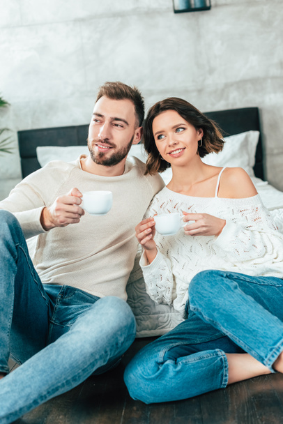 happy man and cheerful woman sitting on floor and holding cups with drinks - Foto, Imagem