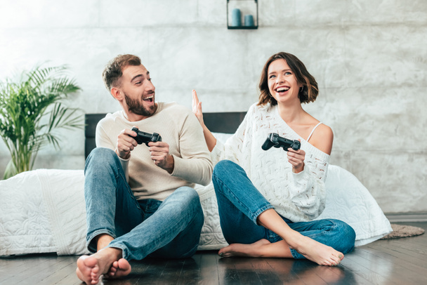surprised man looking at cheerful woman playing video game at home  - Foto, afbeelding