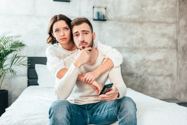 attractive woman hugging upset man sitting on bed and holding remote controller  - Fotó, kép