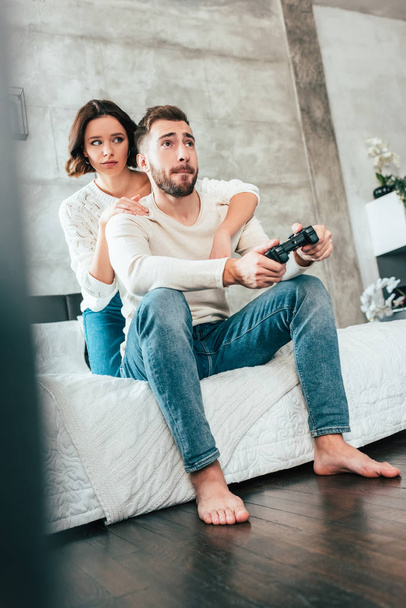 matala kulma näkymä brunette nainen halaus komea mies istuu sängyllä ja pelaa videopeliä
  - Valokuva, kuva