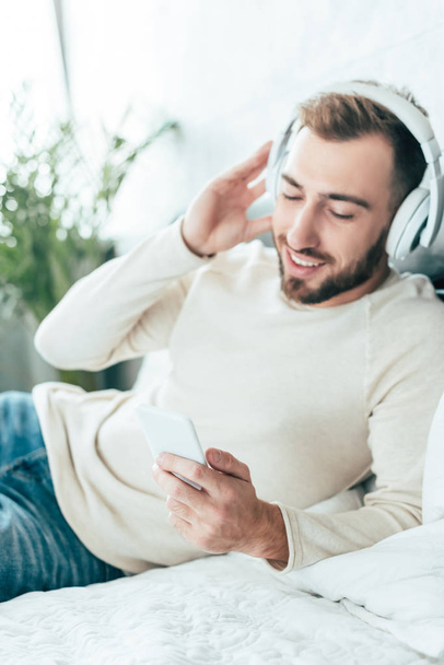 cheerful bearded man using smartphone while listening music in headphones  - Foto, Imagen