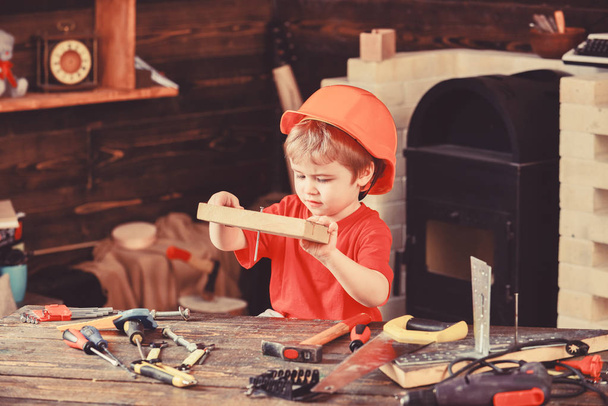 Child in helmet cute playing as builder or repairer, repairing or handcrafting. Handcrafting concept. Toddler on busy face plays at home in workshop. Kid boy hammered nail into wooden board - Фото, изображение