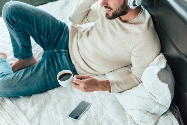 cropped view of cheerful man listening music in headphones while holding cup of coffee in bed  - Zdjęcie, obraz