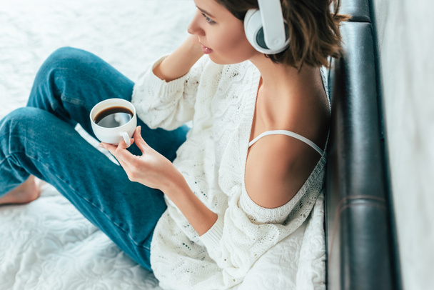 Ansicht einer brünetten Frau mit Kopfhörer, die Tasse hält und Musik hört  - Foto, Bild