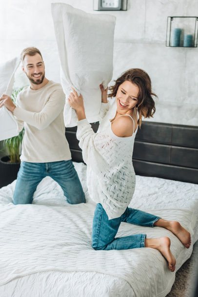 happy bearded man having pillow fight with attractive woman on bed  - Foto, Imagen