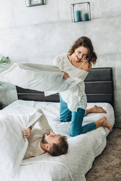 cheerful woman having pillow fight with happy bearded man on bed  - 写真・画像